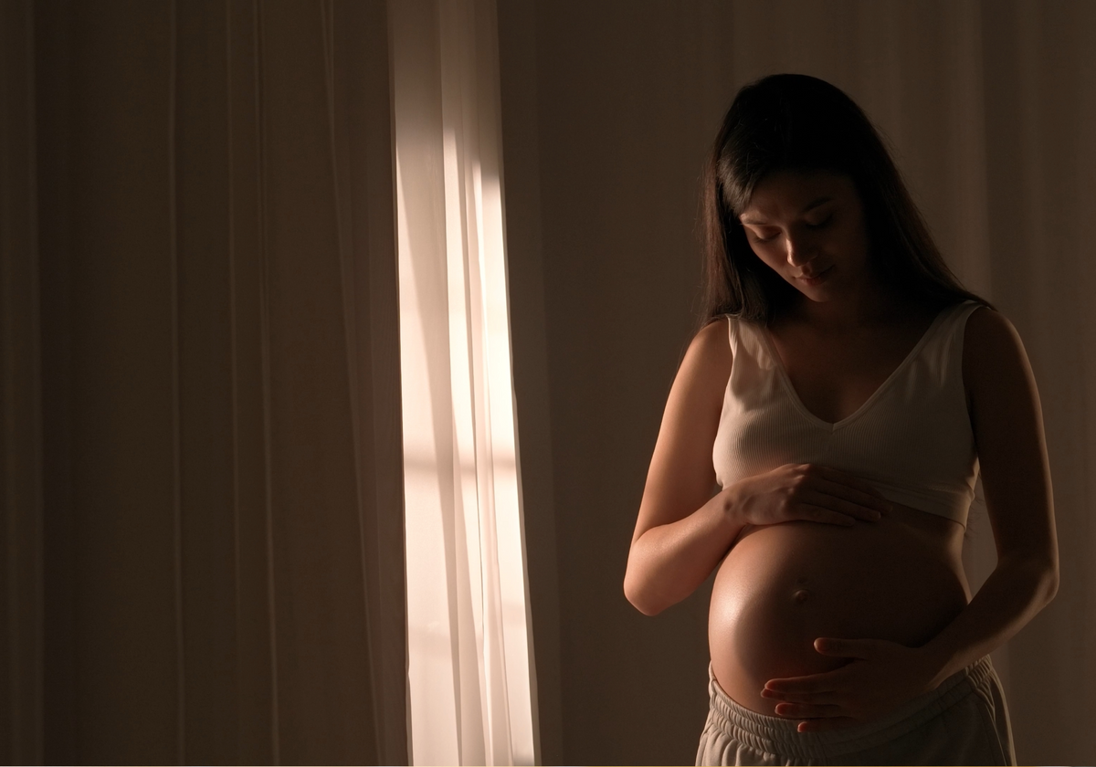 A pregnant woman in a white bra lovingly cradles her baby bump, with soft sunlight filtering through sheer curtains on the left, symbolizing the anticipation and care for the baby that wireless baby monitors full hd systems offer