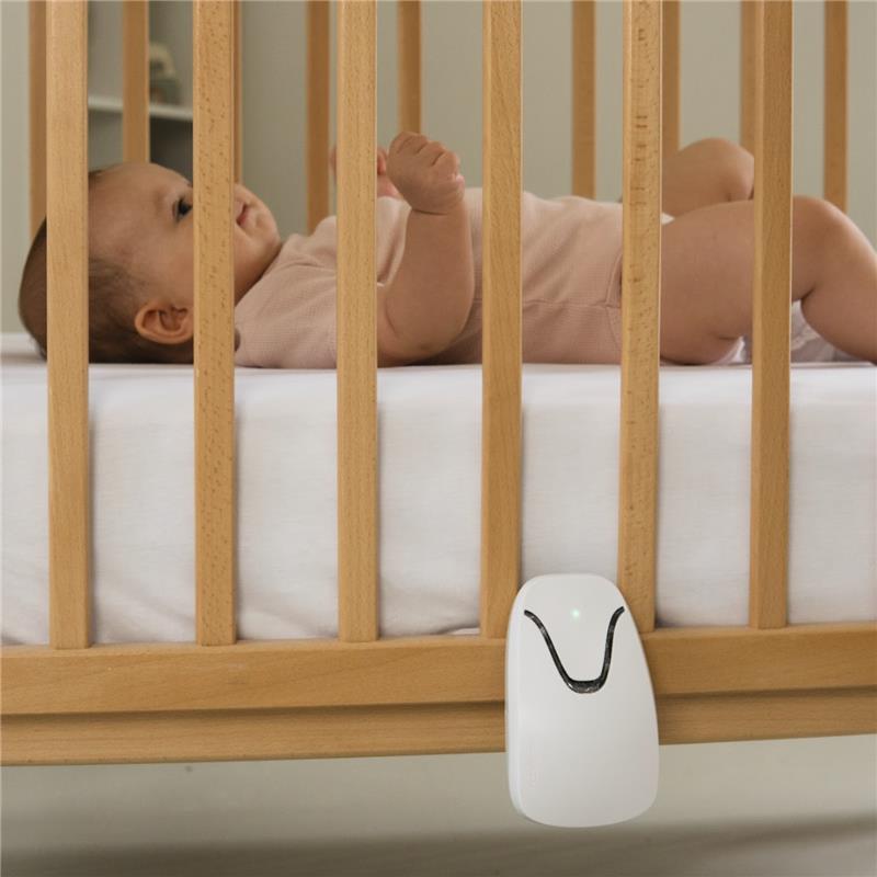 A baby wearing a pink outfit sleeps peacefully in a wooden crib with the white Babysense 7 baby monitor attached to the side, featuring breathing motion detection & safety alerts
