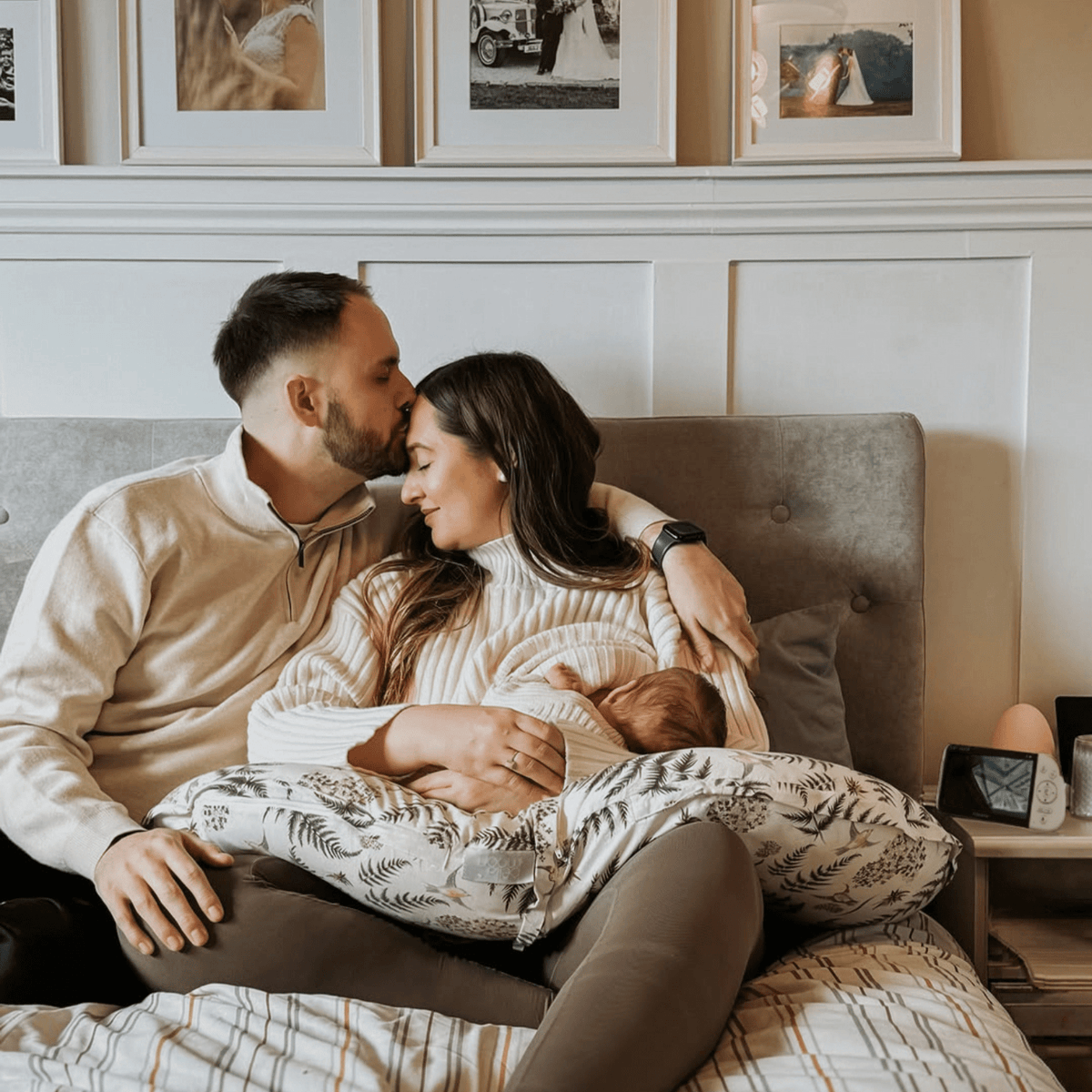 A loving couple on a bed, the father kissing the mother's forehead as she cradles their newborn baby, with an hd video baby monitor on the nightstand next to them