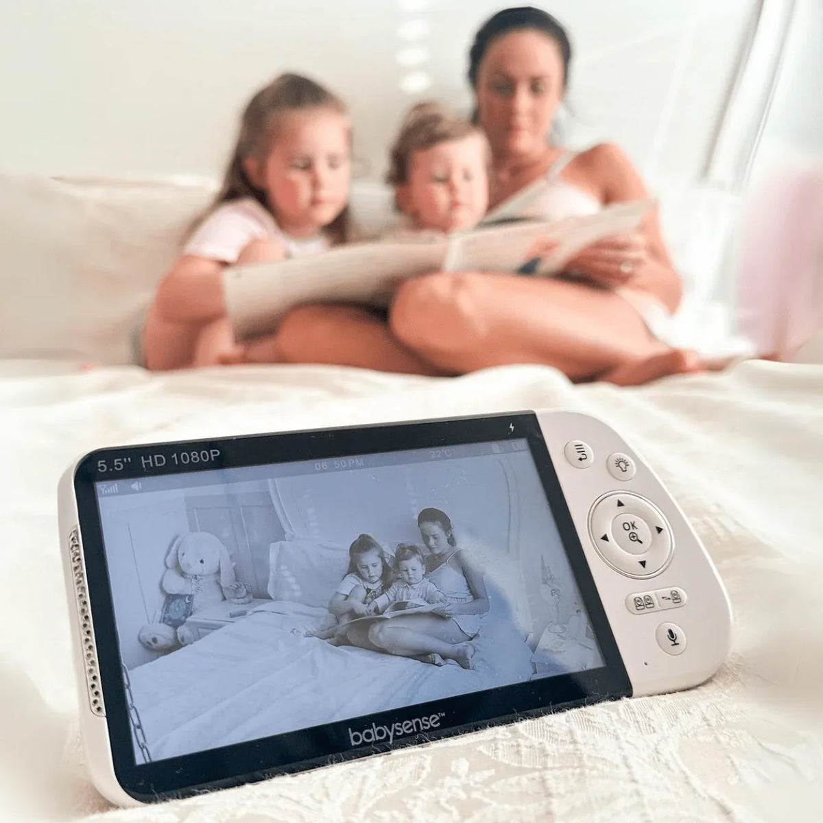 A white Babysense PRO video baby monitor with a 5.5" HD 1080P screen shows a mother reading to her two young daughters in bed, with a wireless HD camera capturing the moment in monochrome