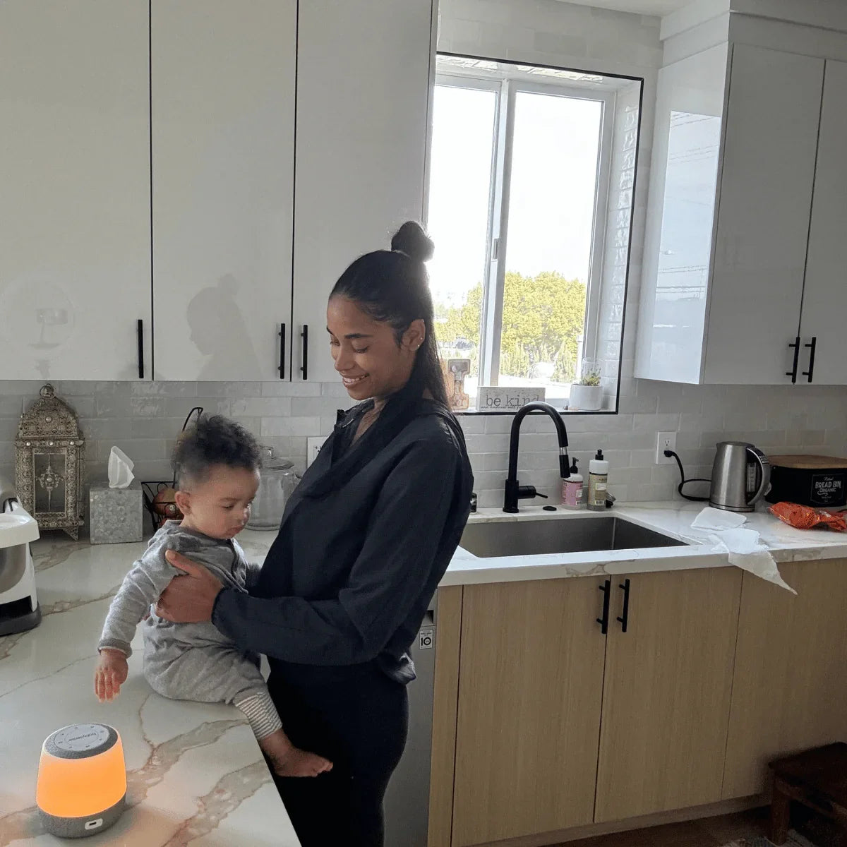 A smiling woman stands in a modern kitchen, holding a baby who sits on the counter next to a glowing Babysense Sleep & Monitor Pack white noise machine, ideal for monitoring with a baby wireless HD camera