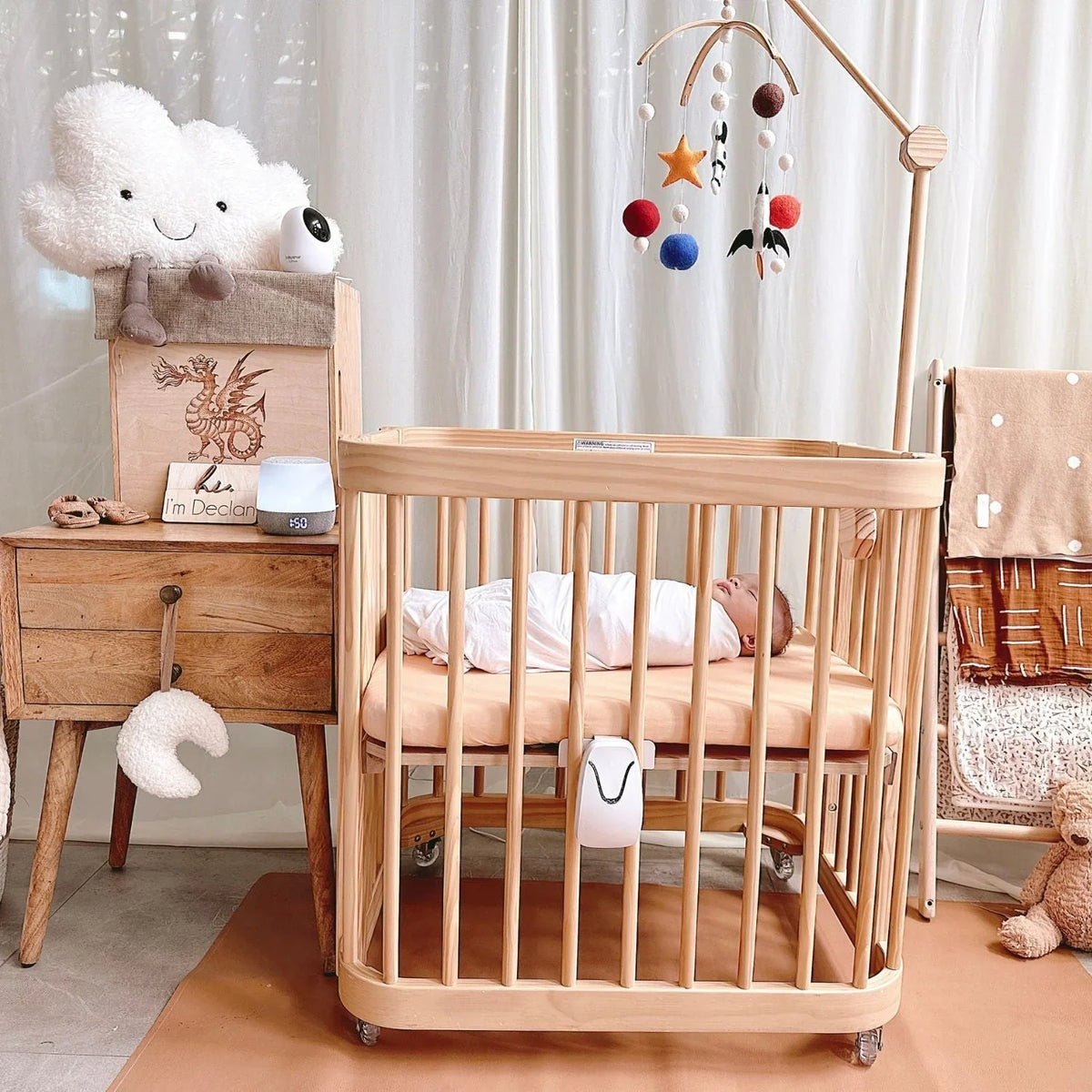 A light wood crib with an infant sleeping inside includes the Babysense Safe Sleep Smart Nursery setup, featuring its video baby monitor, nightlight, and sound machine on a bedside table