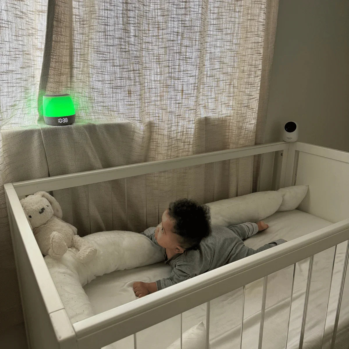 A baby lies on their stomach in a crib with a white stuffed animal as a Babysense FullPeek 3MP HD WiFi baby monitor sits on a nearby shelf, capturing the scene
