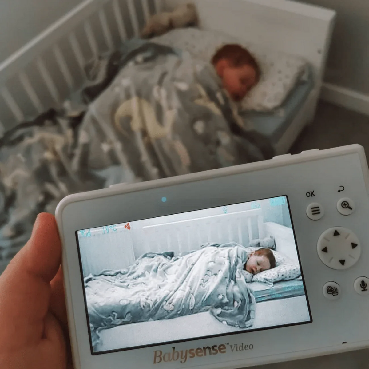 A person holds a white Babysense V43: Video Baby Monitor, showing a clear image of a sleeping baby in a crib on its screen, with the actual baby sleeping in the background, making it an ideal baby monitor with 2 cameras