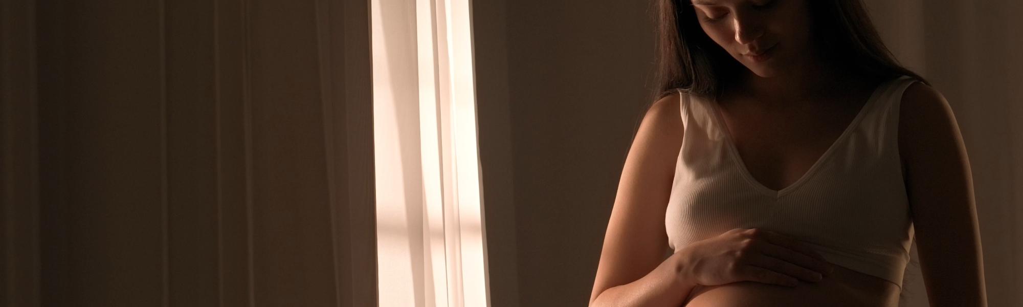 A pregnant woman in a white bra lovingly cradles her baby bump, with soft sunlight filtering through sheer curtains on the left, symbolizing the anticipation and care for the baby that wireless baby monitors full hd systems offer
