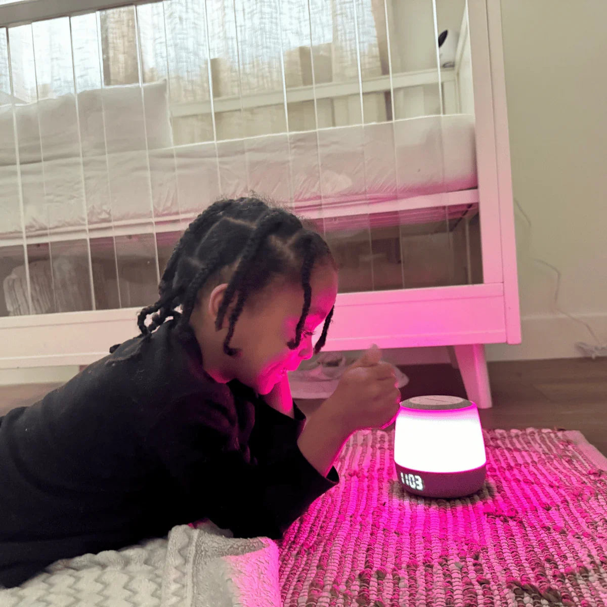 A young child with braided hair lies on their stomach, looking at a Babysense smart baby white noise sound and light machine, which glows pink, on a patterned rug in front of a white crib