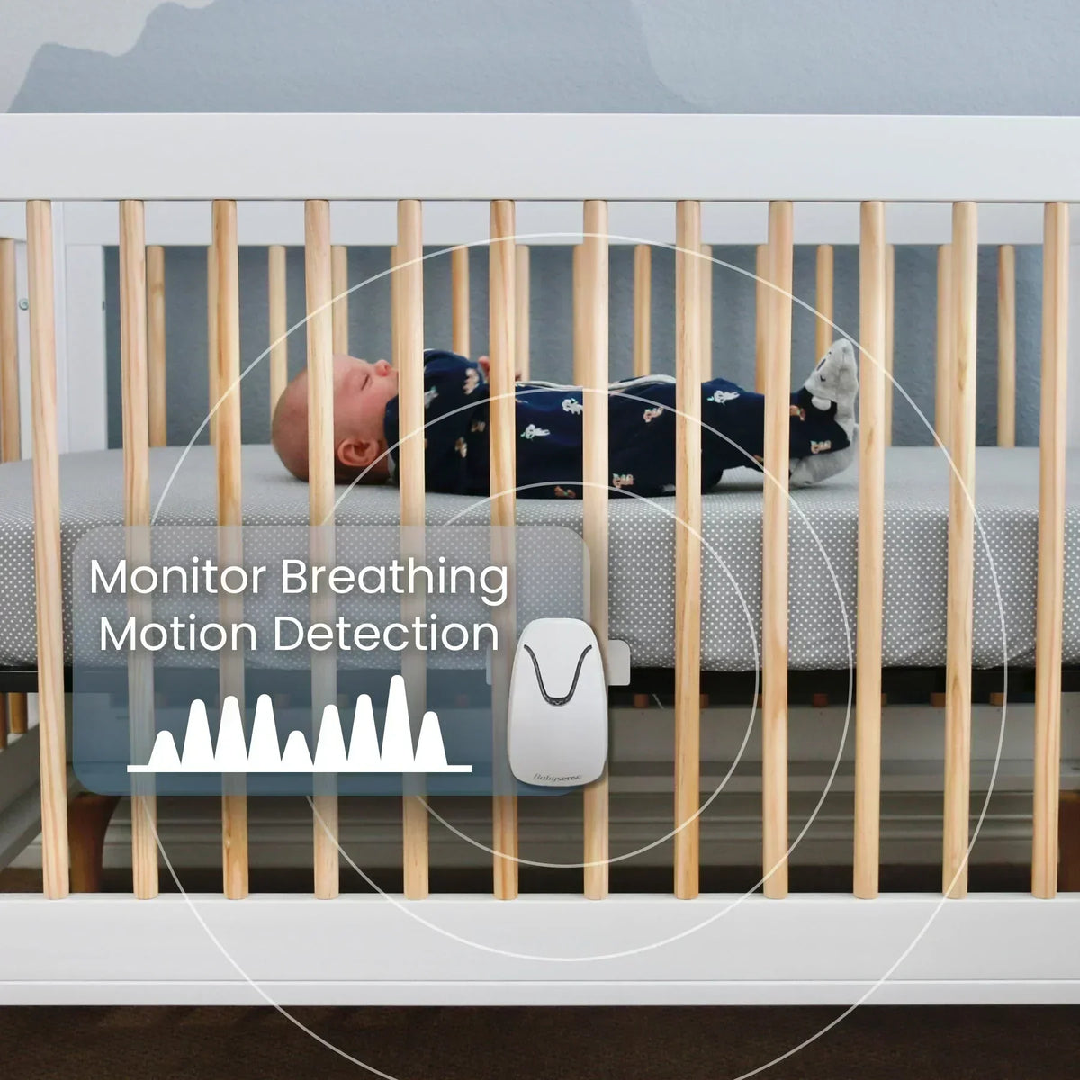 A tiny baby sleeps peacefully in a white crib with natural wood rails, monitored by a white Babysense True Sleep video baby monitor attached to the side with motion detection waves surrounding them