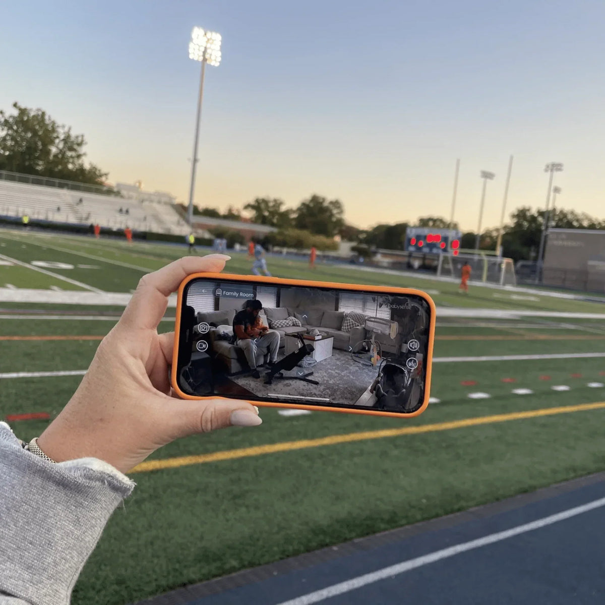 A hand holds a phone displaying the Babysense app, showing a man and baby in a family room, at a sports field at sunset, highlighting the baby monitor with sleep tracking