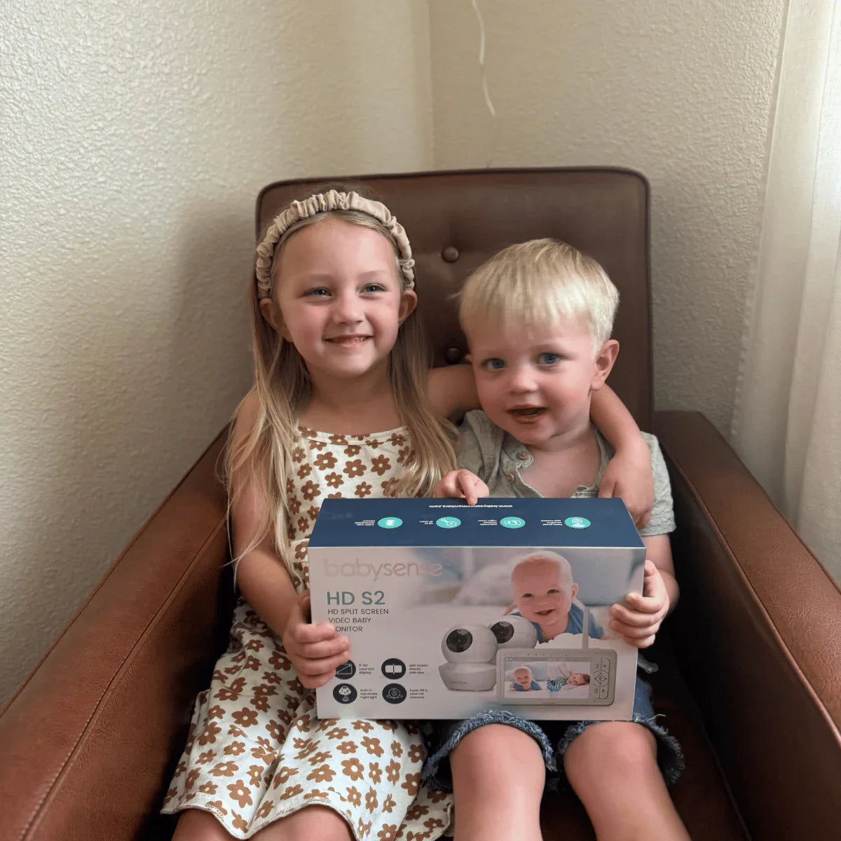 Two smiling young children sit in a brown chair, holding a Babysense HD S2 box for a video baby monitor with both wifi and non wifi capabilities