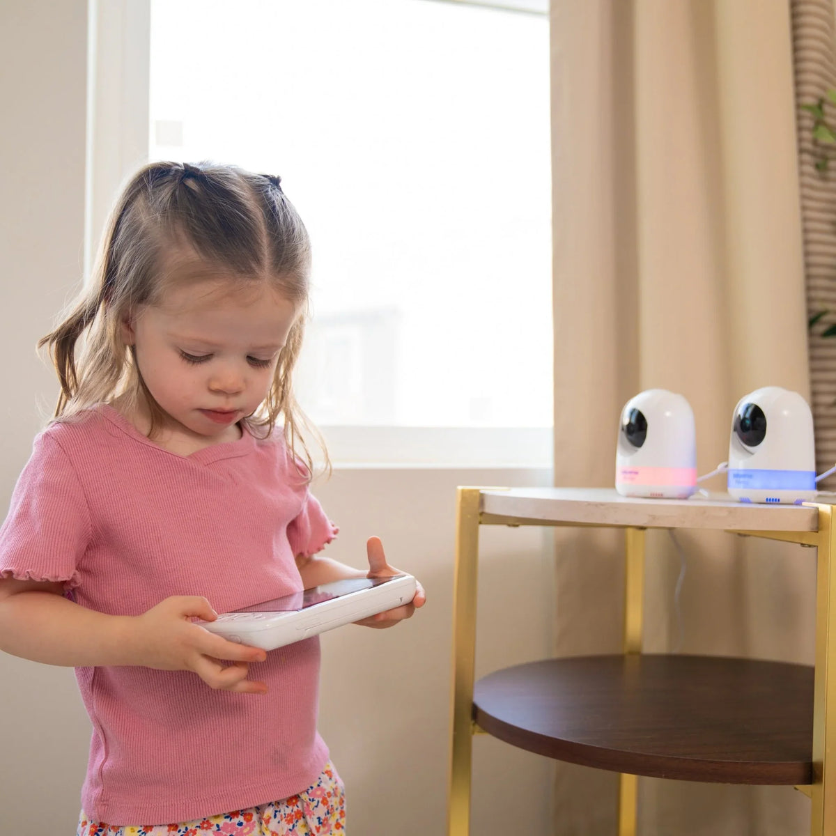 A young girl in a pink shirt holds the Babysense Prisma video baby monitor handheld screen while two white baby wireless HD camera units with glowing bases sit on a table