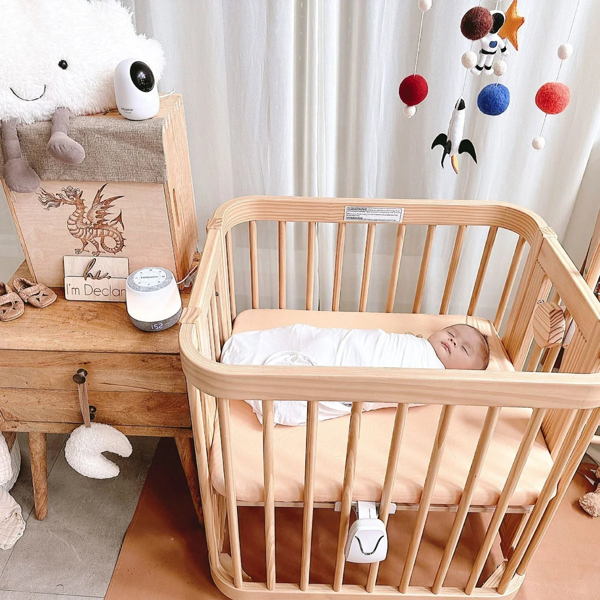A Babysense smart nursery camera, a video baby monitor, and a white sound machine sit on a wooden chest and nightstand beside a baby sleeping in a crib