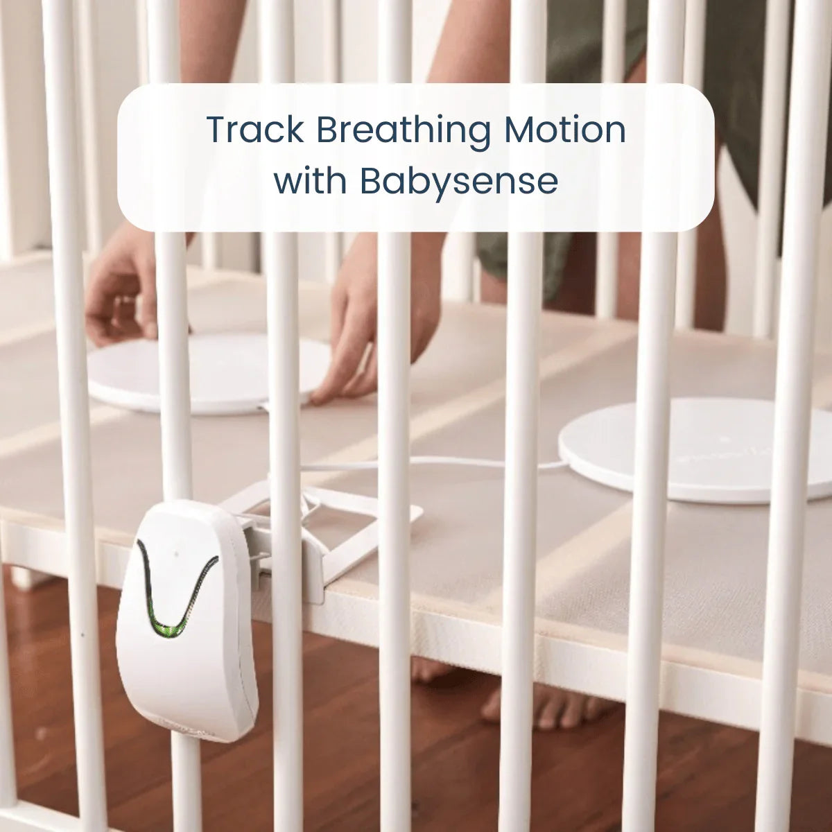 A person's hands arrange two white circular sensors, part of the Babysense Safe Sleep Smart Nursery, in a white crib, designed to track breathing motion with safety alerts