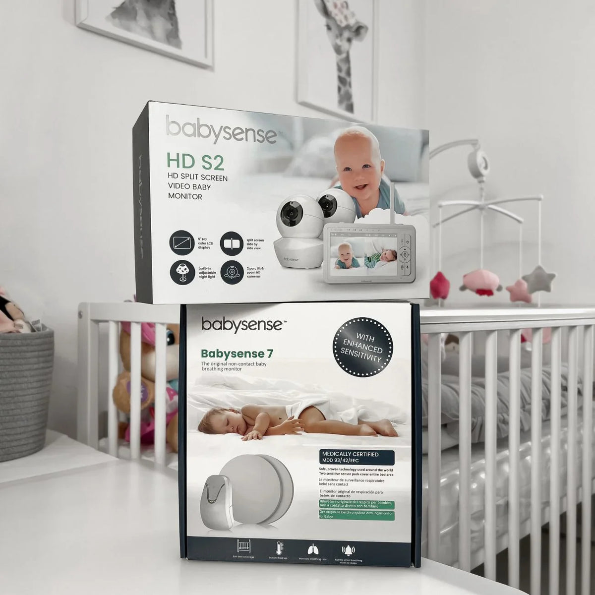 Babysense True Sleep: Video Baby Monitor with Breathing Motion Detection & Safety Alerts is seen in its packaging, with a white video baby monitor box stacked on top of a gray baby sensor breathing monitor box, both prominently featuring babies and product details on a white table next to a crib