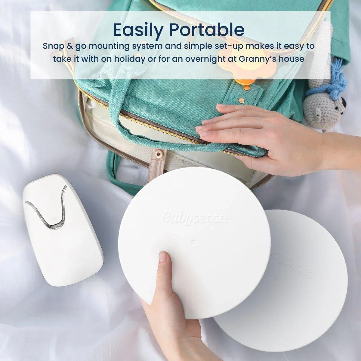 A cropped close-up shows a white Babysense 7 video baby monitor receiver and two circular sensor pads being packed into a green diaper bag, highlighting the easy portability for travel