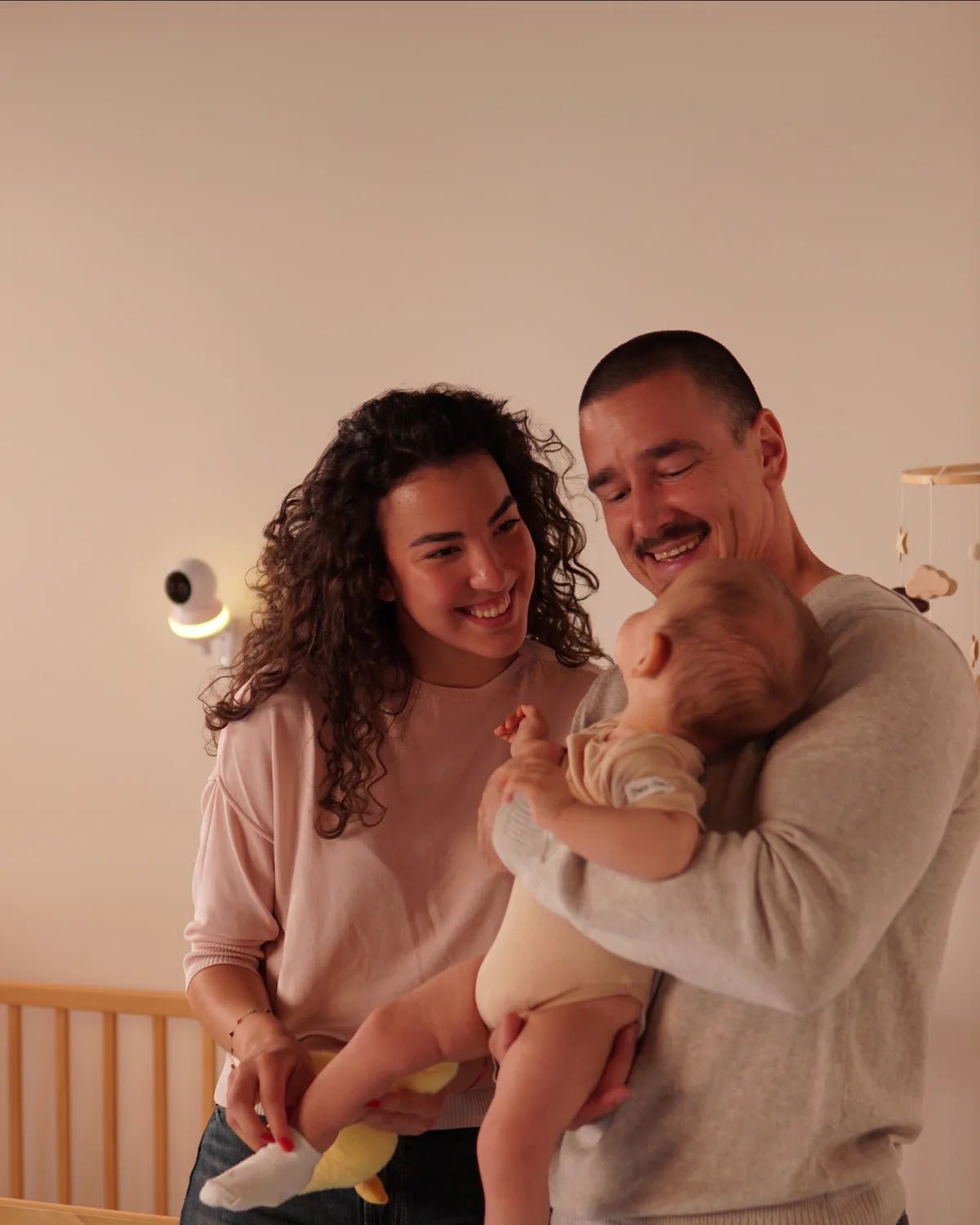 Parents smiling at their baby in the nursery, with the Babysense camera glowing on the wall